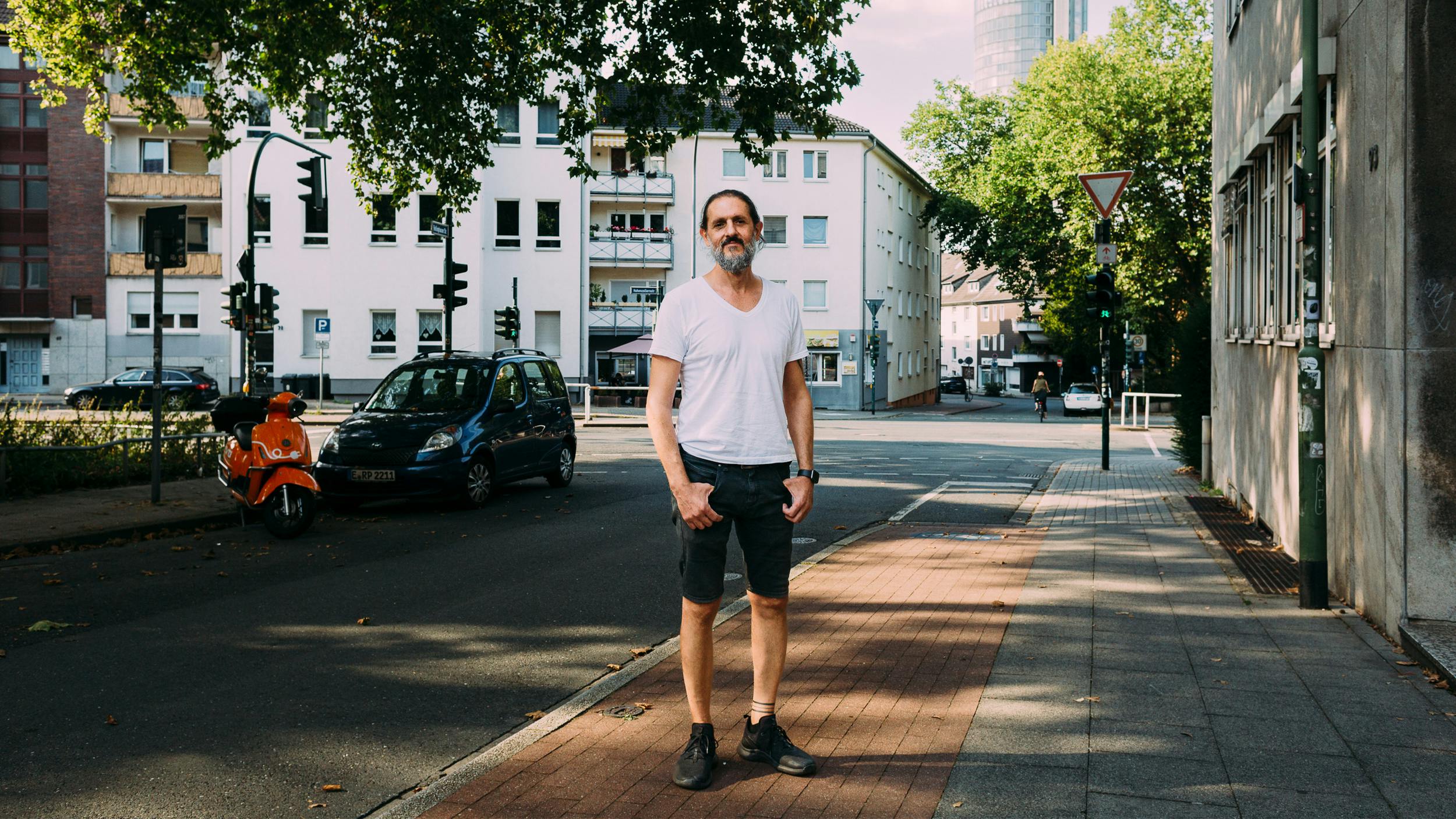 Saad steht auf einem Bürgersteig in der Essener Innenstadt. Er trägt ein weißes T-Shirt, eine kurze schwarze Jeanshose und lächelt in die Kamera. Im Hintergrund sind Wohnhäuser und ein Hochhaus zu erkennen.