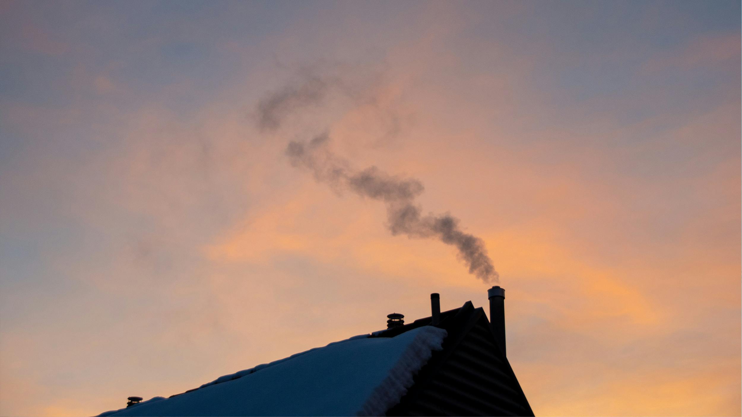 Ein schneebedecktes Dach bei Sonnenuntergang. Aus dem Schornstein steigt Rauch auf.