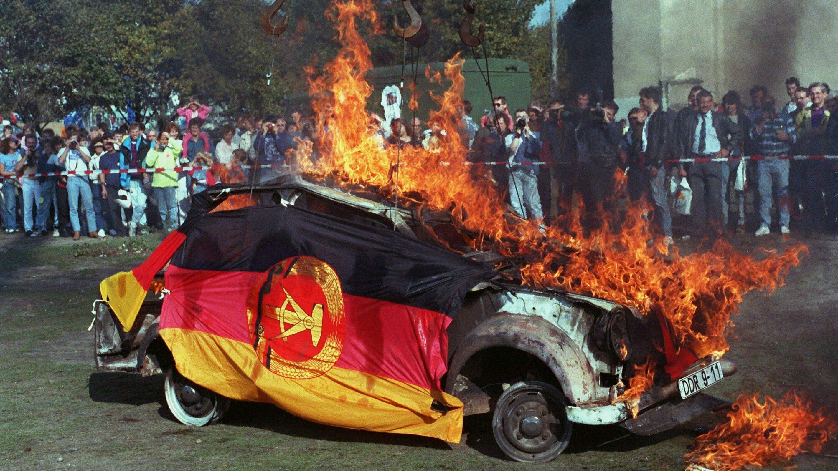 Ein brennender Trabi. Er ist in eine DDR-Fahne gehüllt.