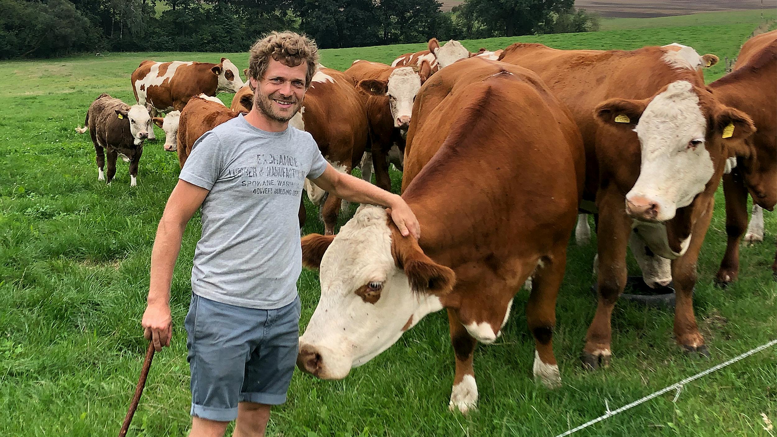 Landwirt Christoph steht neben seinen Kühen. Er blickt in die Kamera und streichelt eine Kuh am Kopf.