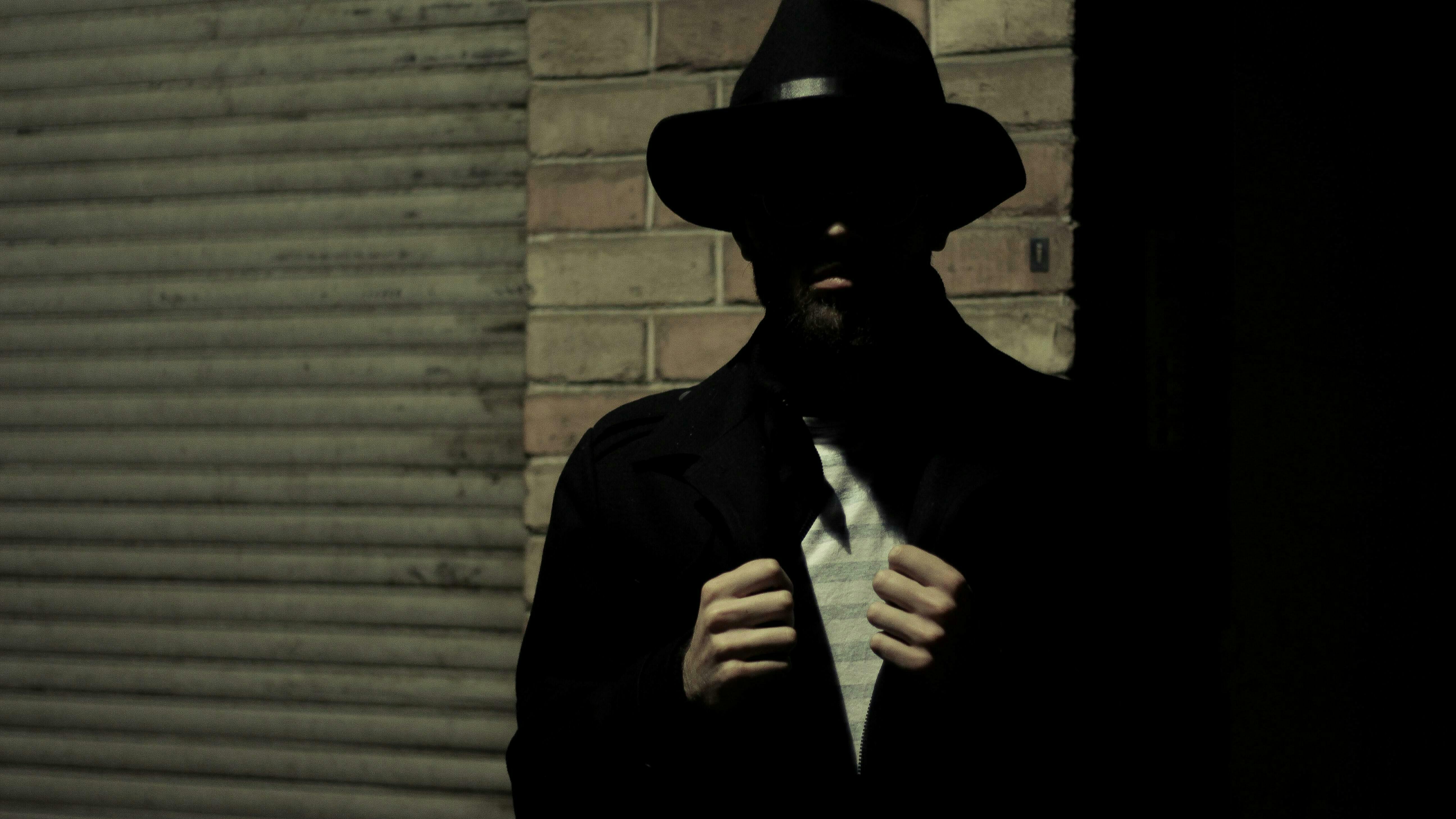 Ein Mann steht vor einer Wand. Sein Gesicht liegt im Schatten. Er trägt einen Hut.