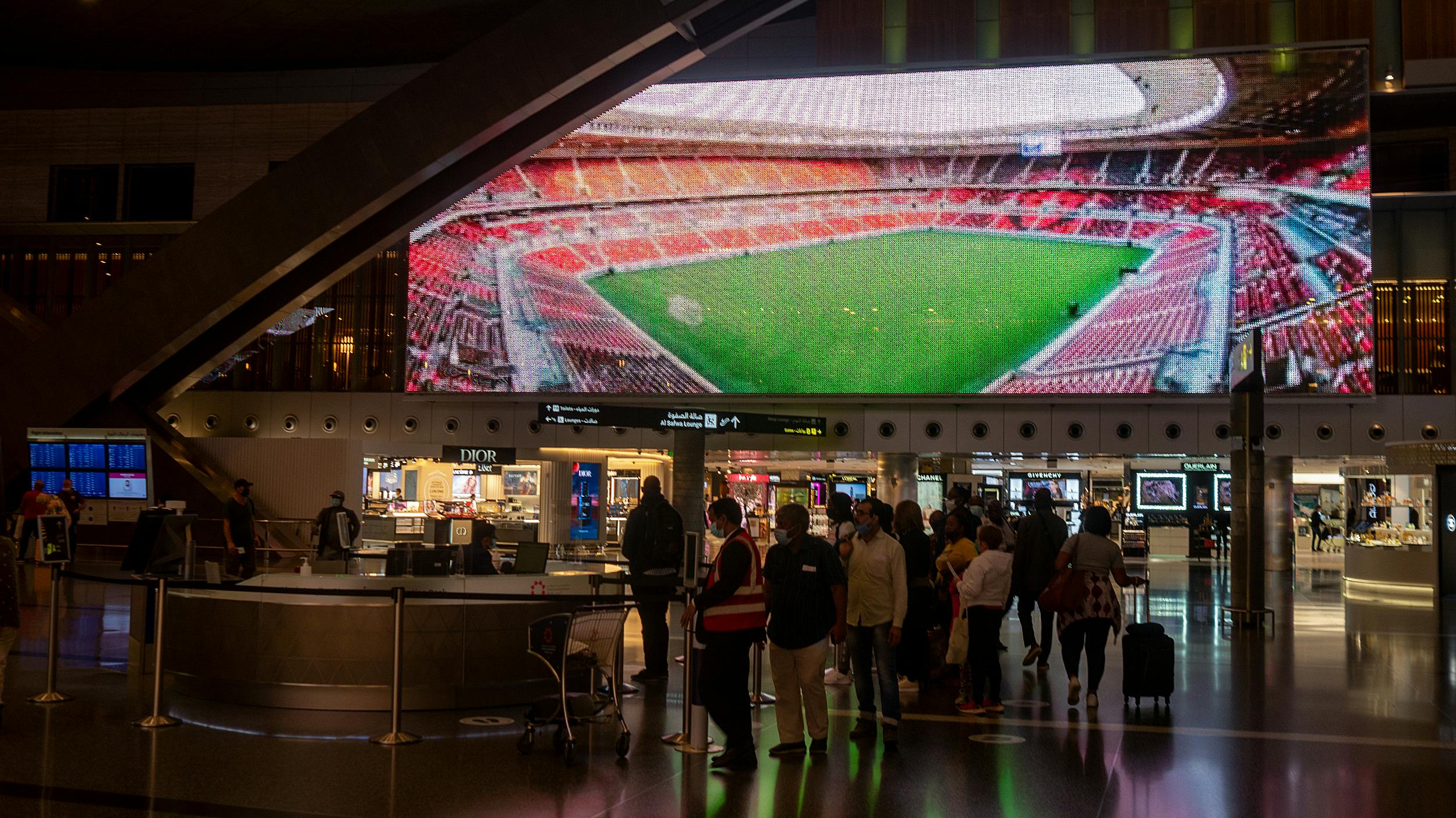 Menschen stehen in einem Flughafen Schlange. Hinter ihnen leuchtet das Bild eines Stadions auf einer LED-Wand.