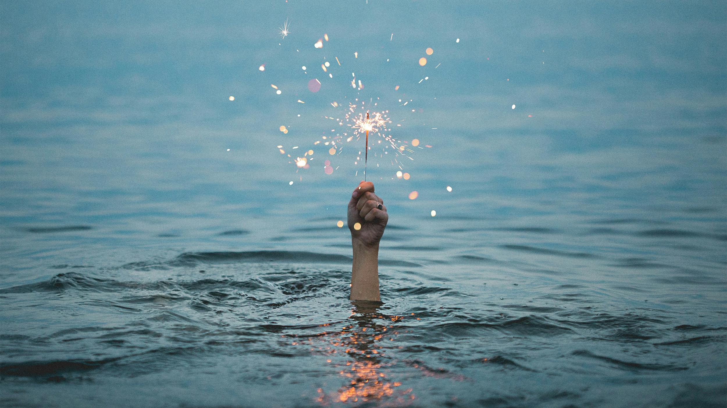 Eine Hand ragt aus tiefem Wasser. Sie hält eine Wunderkerze.