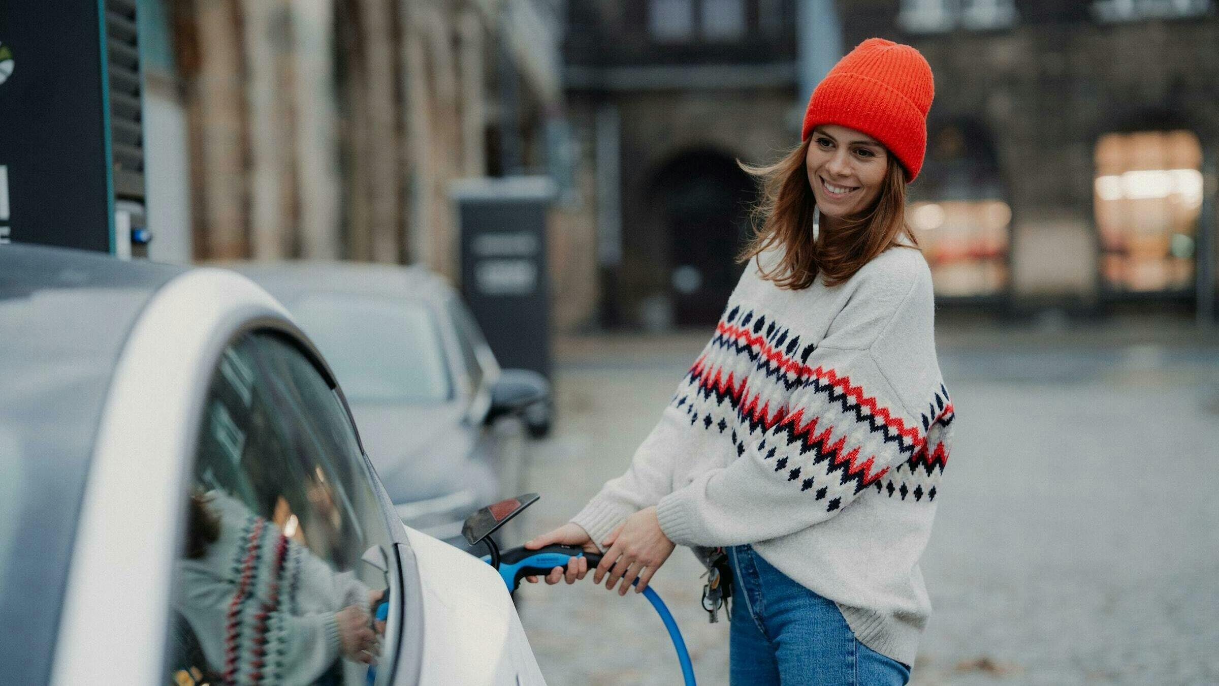 Eine Frau mit einer roten Mütze und Norwegerpullover tankt ihr elektronisches Auto mit Strom.