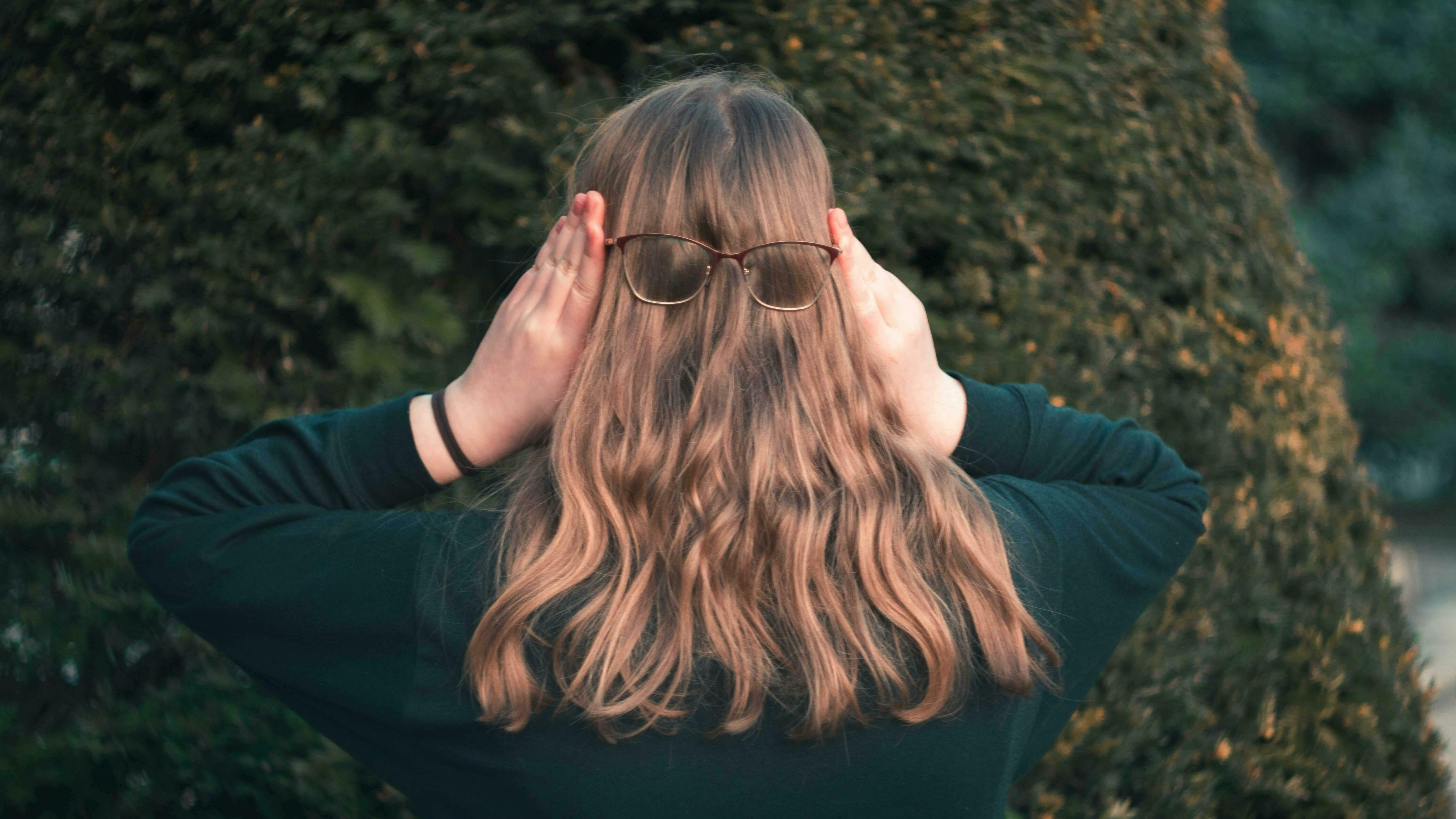 Man sieht eine junge Frau von hinten mit langem, rotbraunen Haar, die sich auf ihr Haar eine Brille setzt.