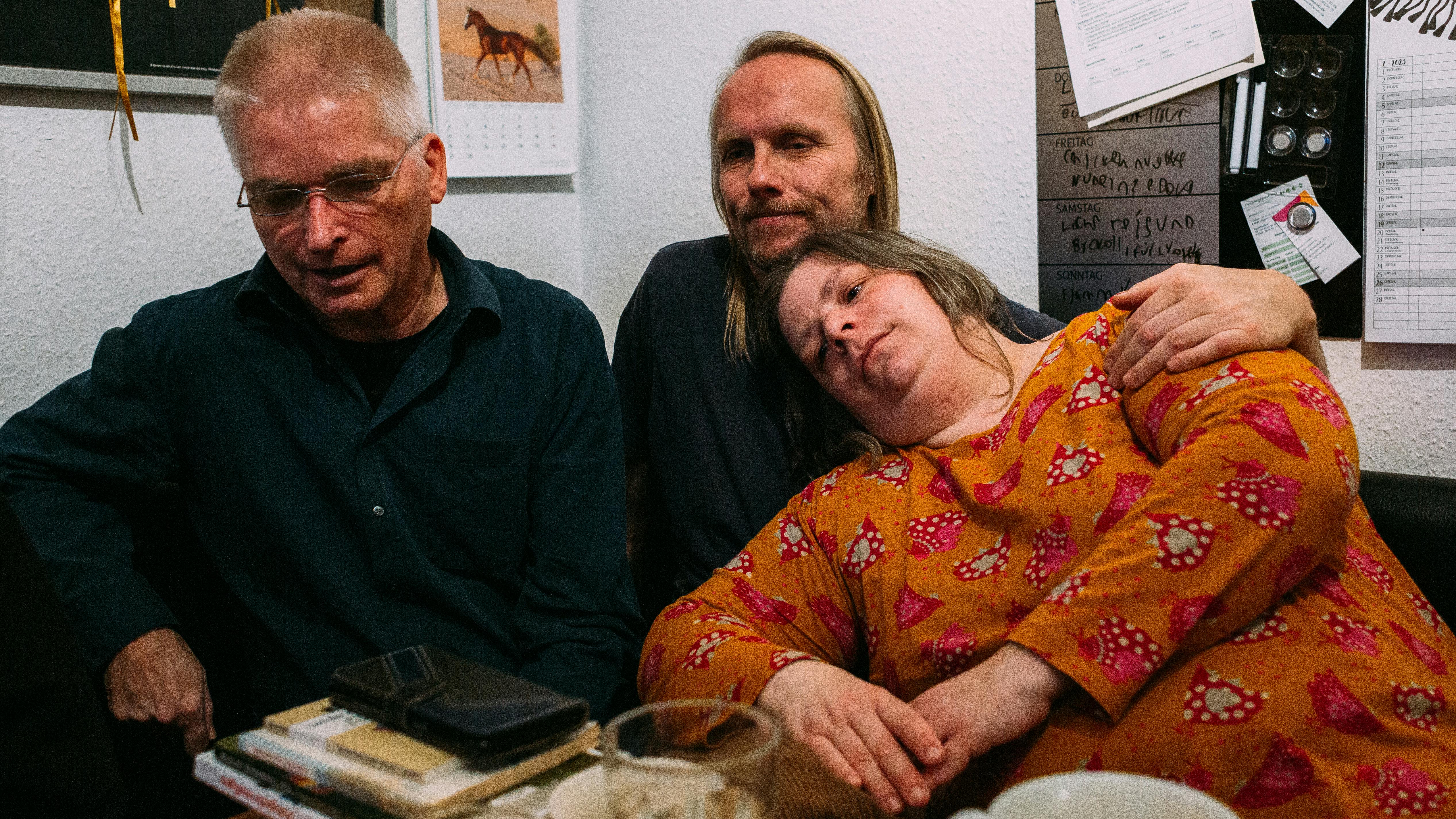 Das Foto zeigt drei Personen auf einer Couch nebeneinander. Rechts ist Susa, in einem orangen Shirt. Sie lehnt an Arnes Brust, der in der Mitte sitzt. Neben ihm auf der linken Seite ist Rainer. Er sieht nach unten und hat den Mund zum Sprechen offen.