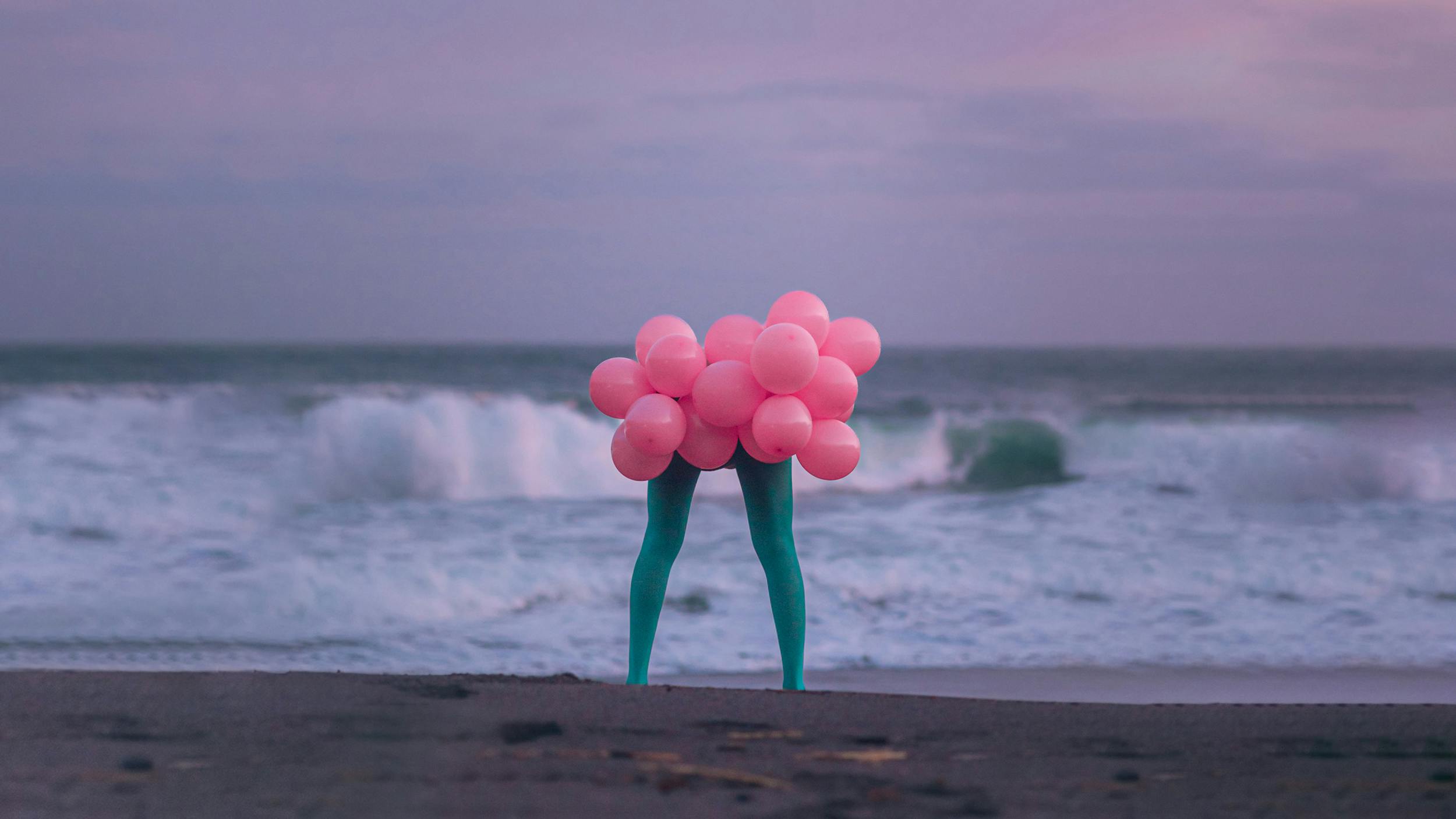 Fotomontage: Ein paar Beine in grünen Leggings steht am Strand. Dort, wo der Oberkörper anfangen würde, befindet sich ein Haufen rosafarbener Luftballons.