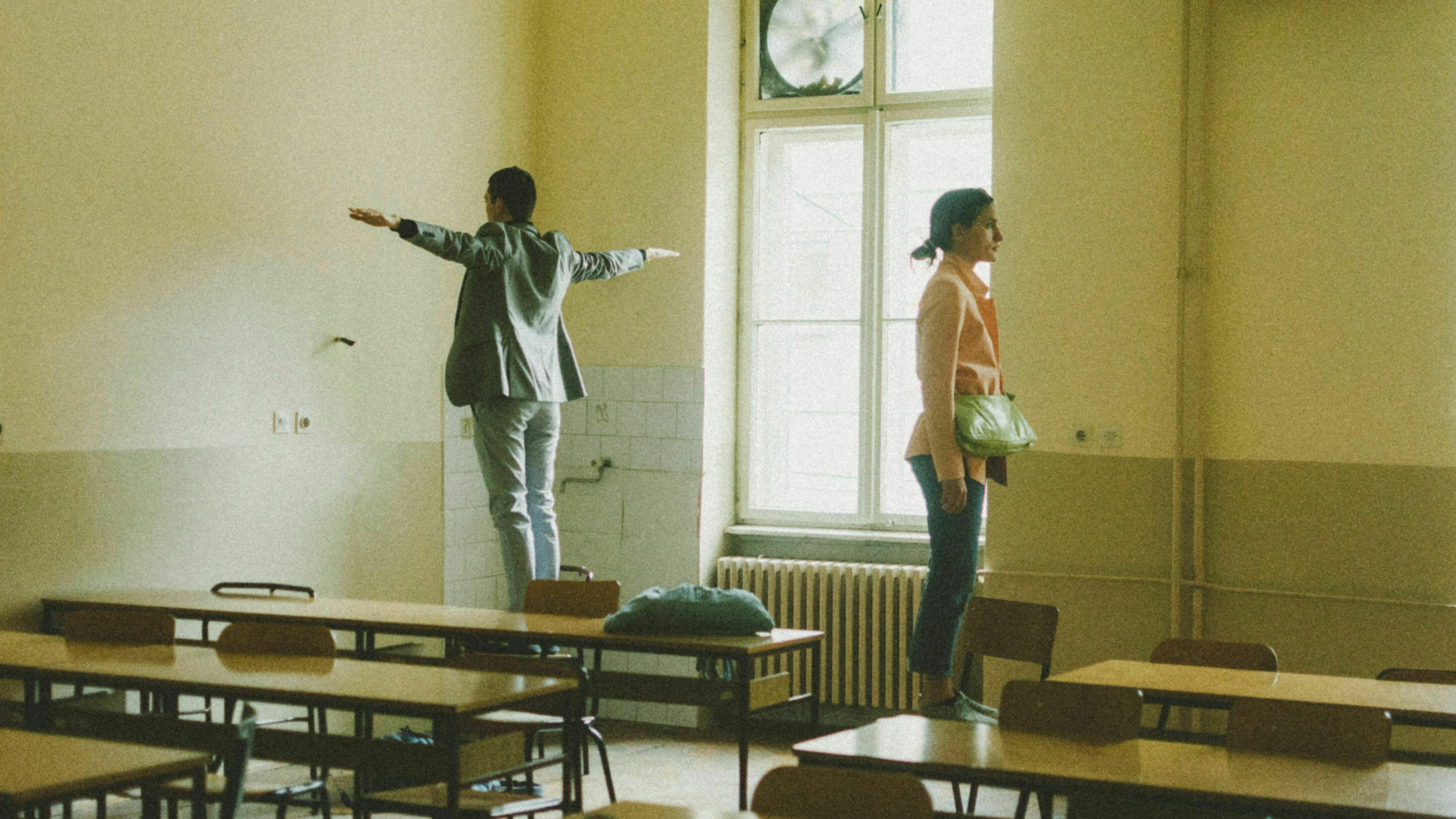 Ein Mann und eine Frau stehen in einem Klassenraum auf zwei Stühlen und gucken die Wände an. Der Mann breitet seine Arme aus. 