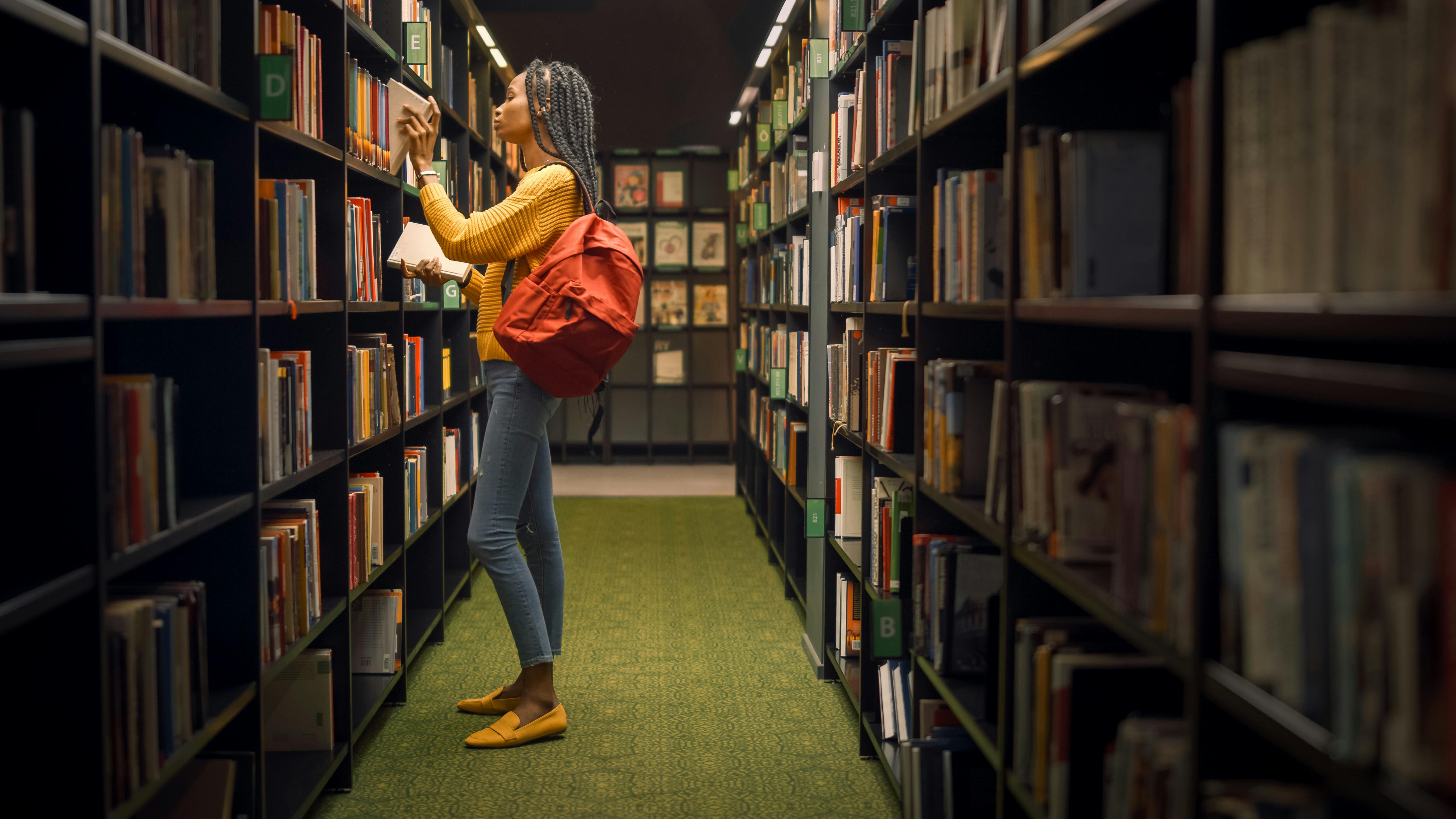 Wir sehen eine schwarze Frau, die in einem Gang in einer Bibliothek steht