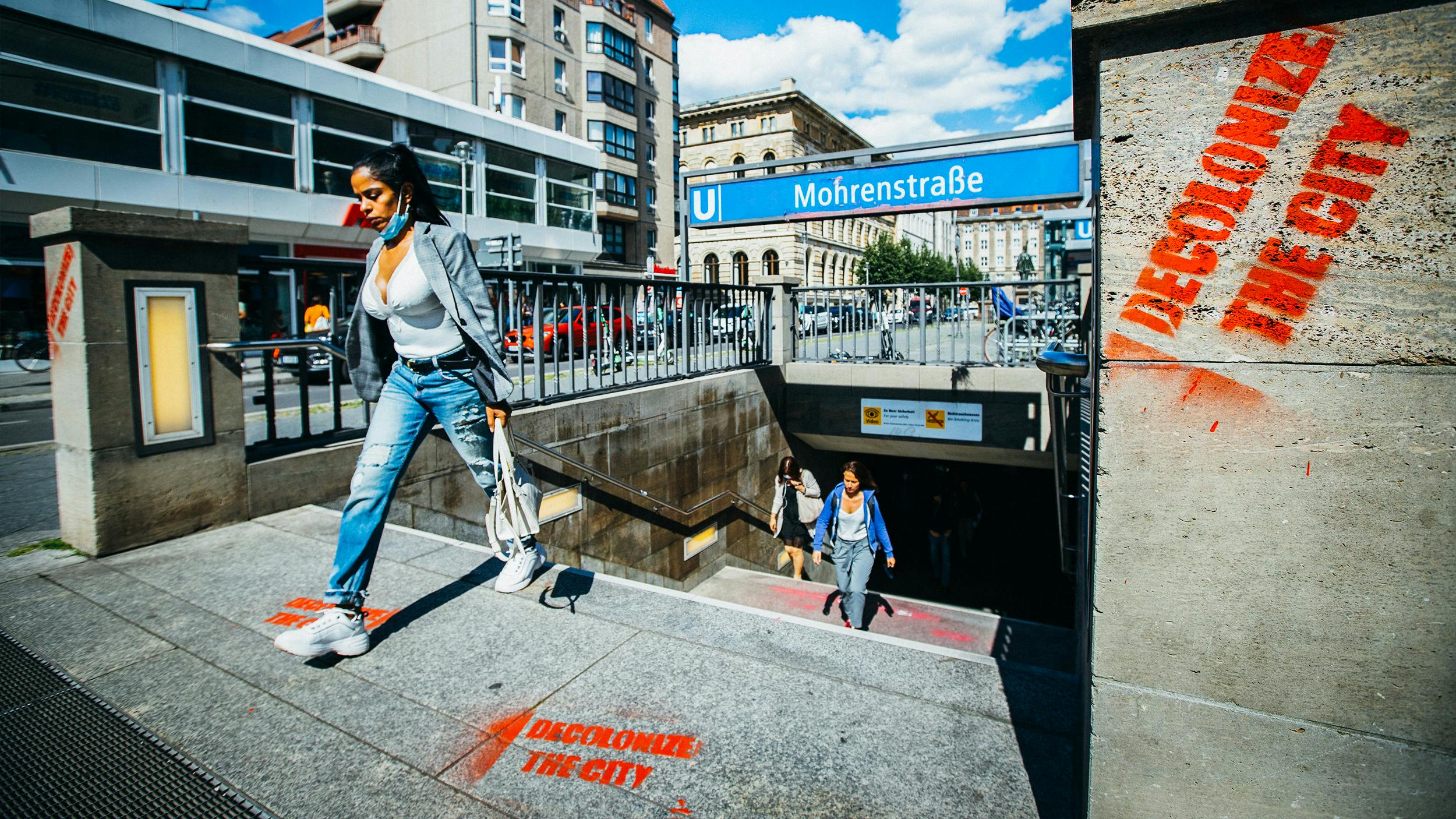 Eine Frau läuft aus der U-Bahn-Station Mohrenstraße. Graffiti in roten Farben proklamieren: „Decolonialize the city“