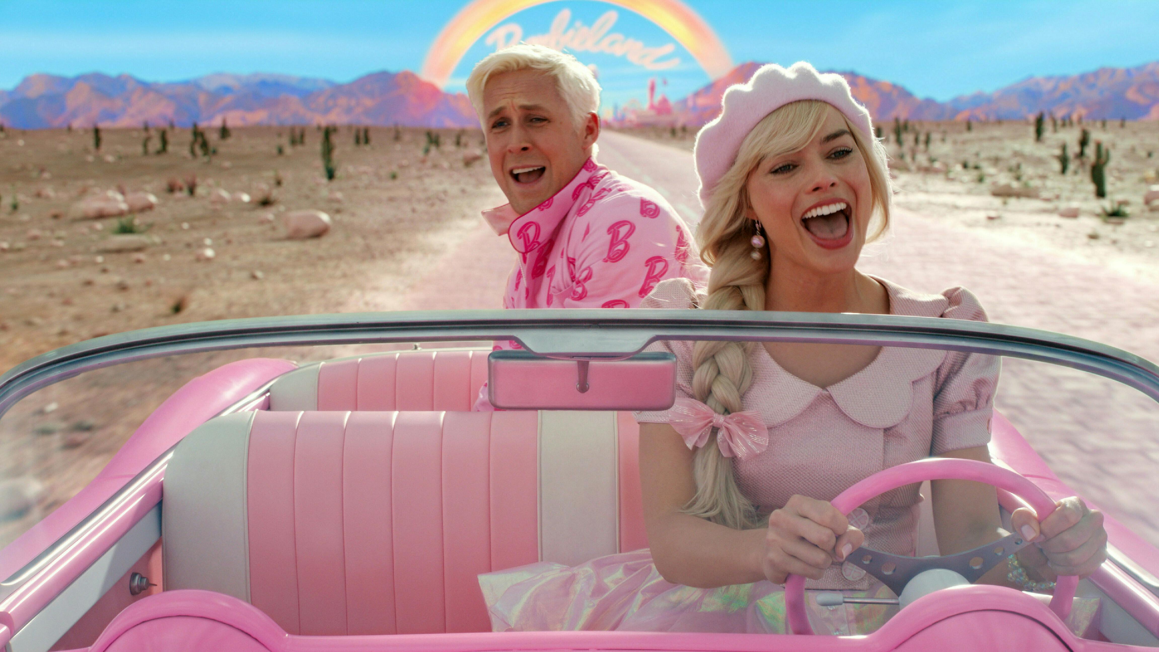 Man sieht die Schauspieler:innen Margot Robbie und Ryan Gosling in der Barbie-Filmkulisse: Barbie sitzt im Auto und Ken auf der Rückbank.