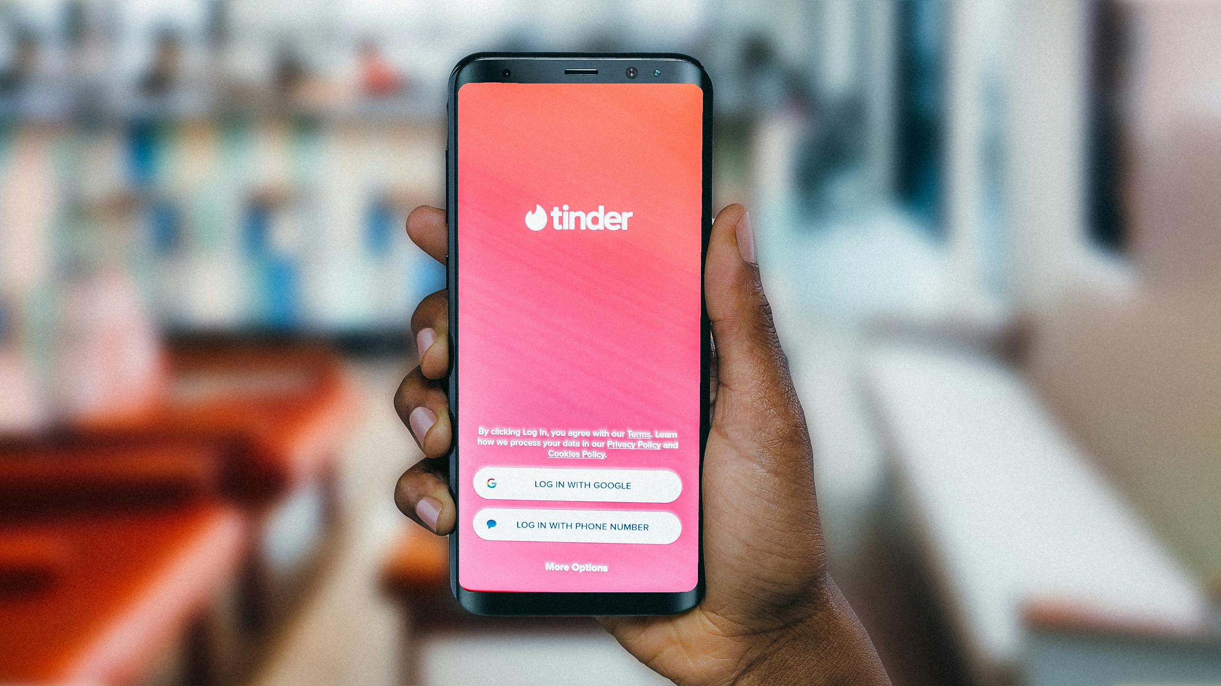 Man sieht die Hand einer person of colour, die ein Handy in der Hand hält, auf der man das Startbild der Online-Datingplattform Tiinder sieht.