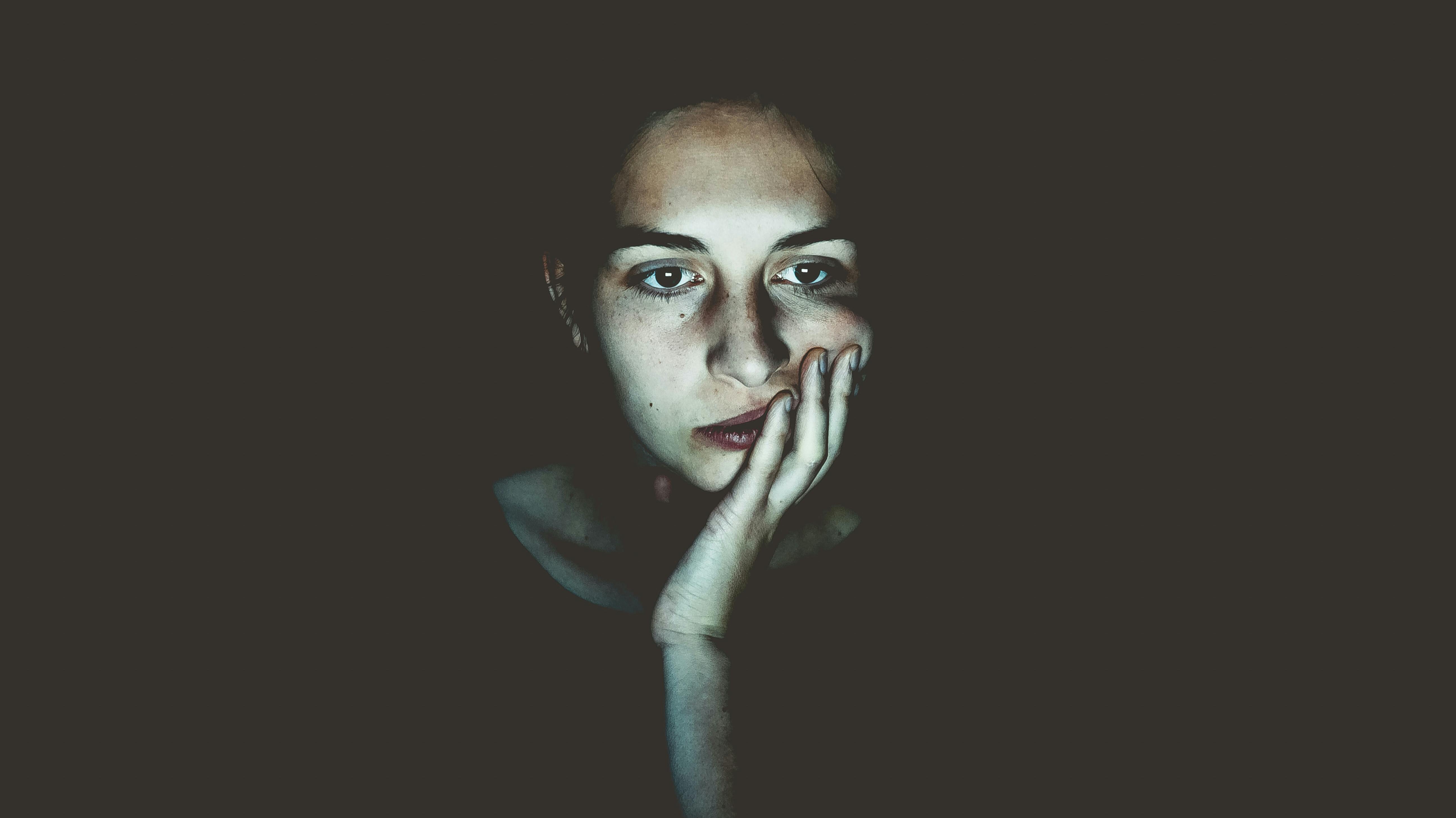 Eine junge Frau sitzt mit müdem Blick vor einem dunklen Hintergrund. 