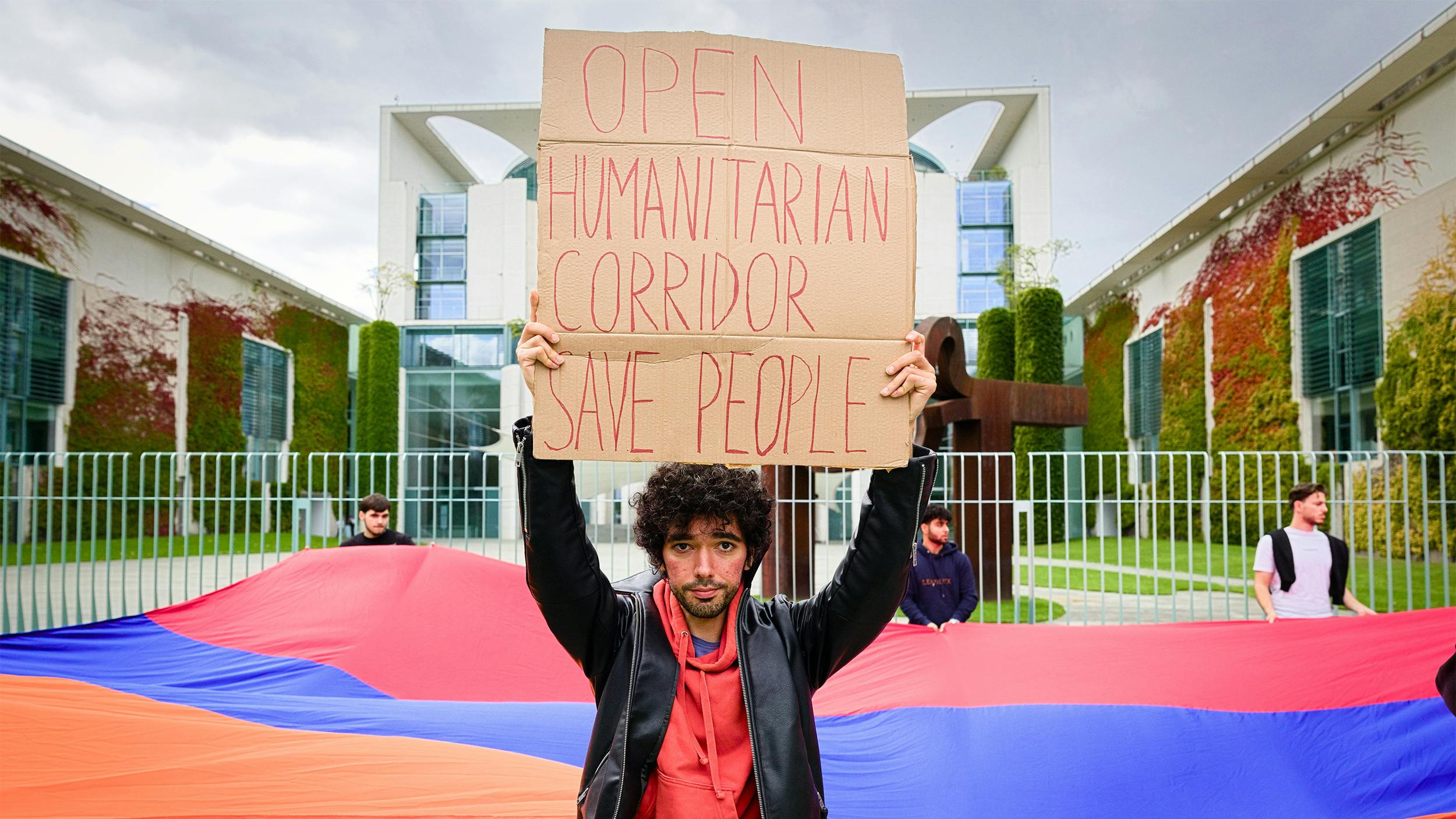 Ein junger Mann hält ein Schild vor dem Kanlzeramt. Darauf steht: „Open humanitarian corridor, save people“-