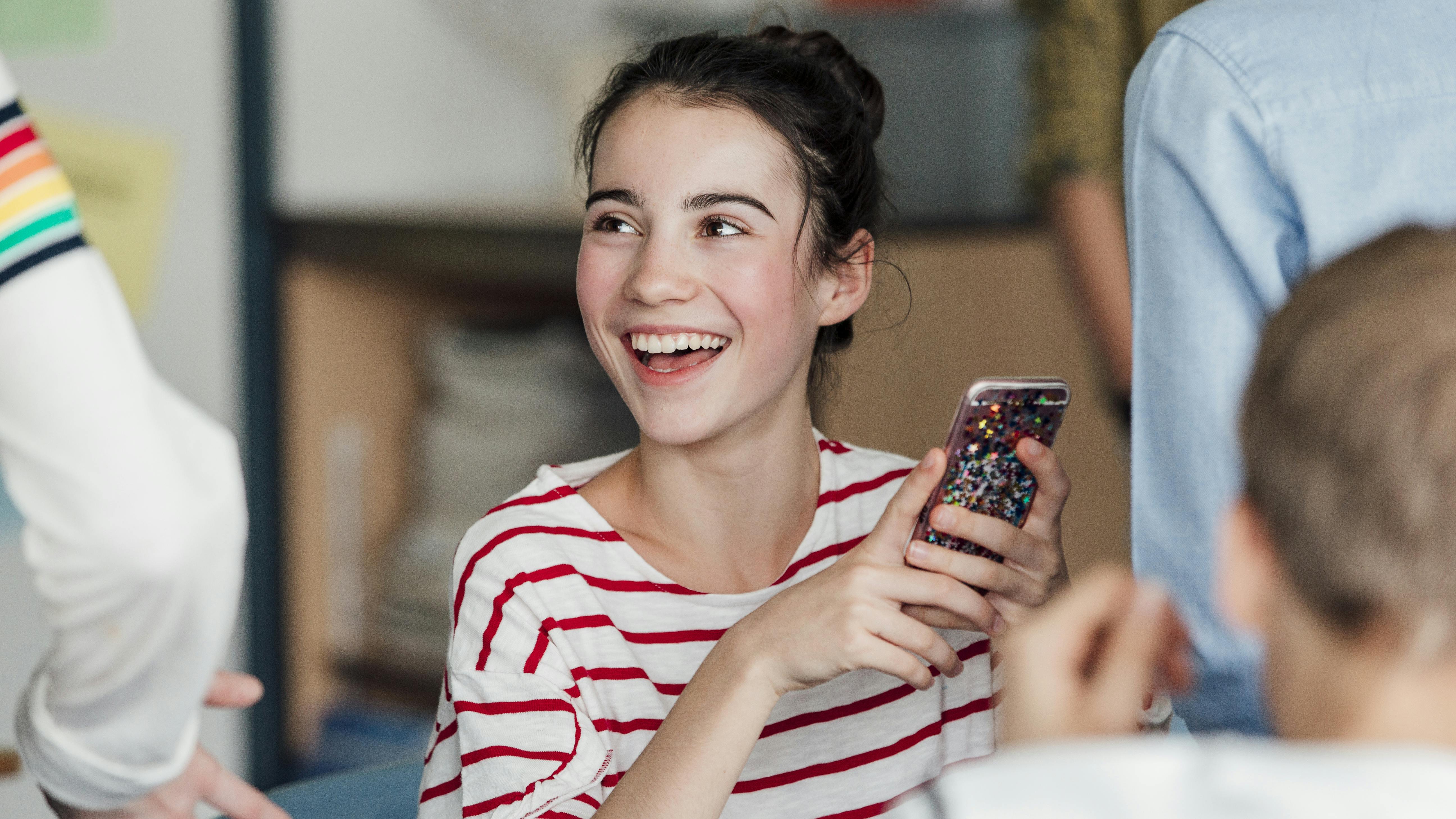 Ein Mädchen sitzt in der Schule vor ihrem Tisch mit dem Smartphone in der Hand. Sie lacht. 