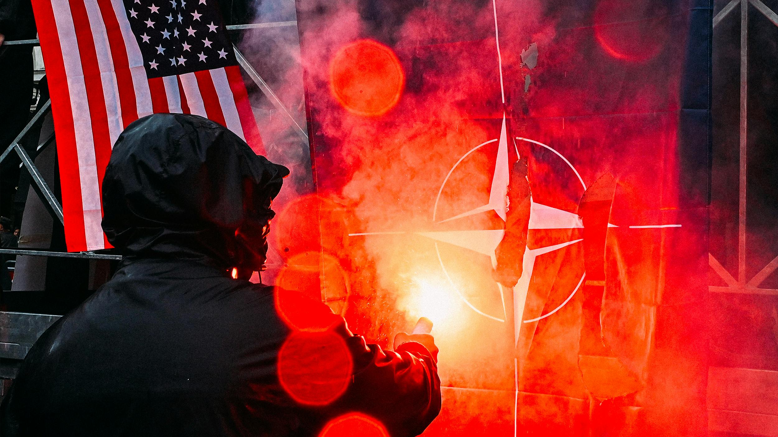 Ein schwarz gekleideter Mensch verbrennt eine NATO Flagge auf einer Demonstration. Im Hintergrund ist eine Amerikanische Flagge zu sehen.