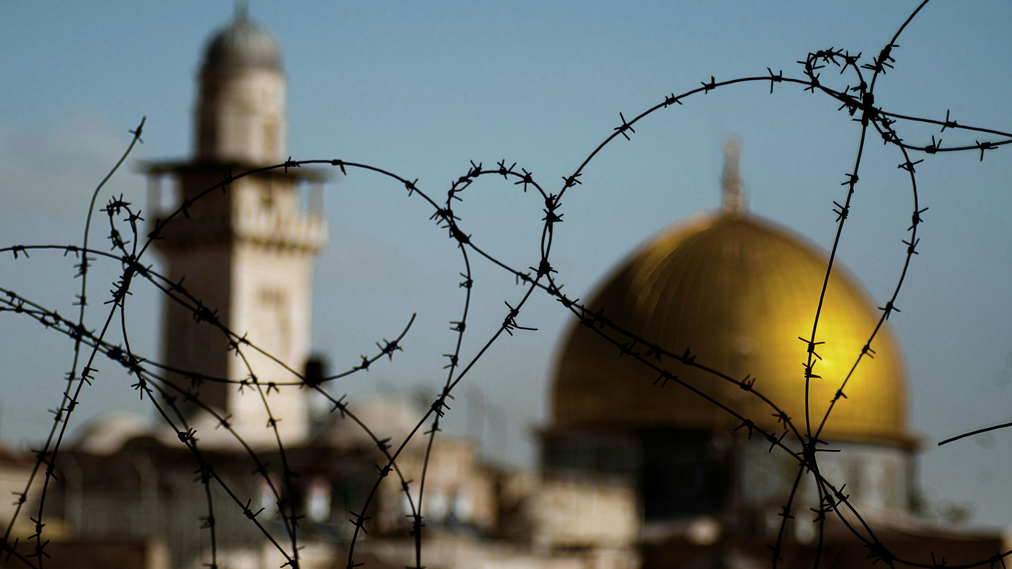 Blick auf die goldene Kuppel des Felsendoms in Jerusalem, unscharf im Hintergrund, durch Stacheldraht scharf im Vordergrund. Das Bild fängt das Konzept des Konflikts und der Hoffnung auf Frieden im Nahen Osten ein.