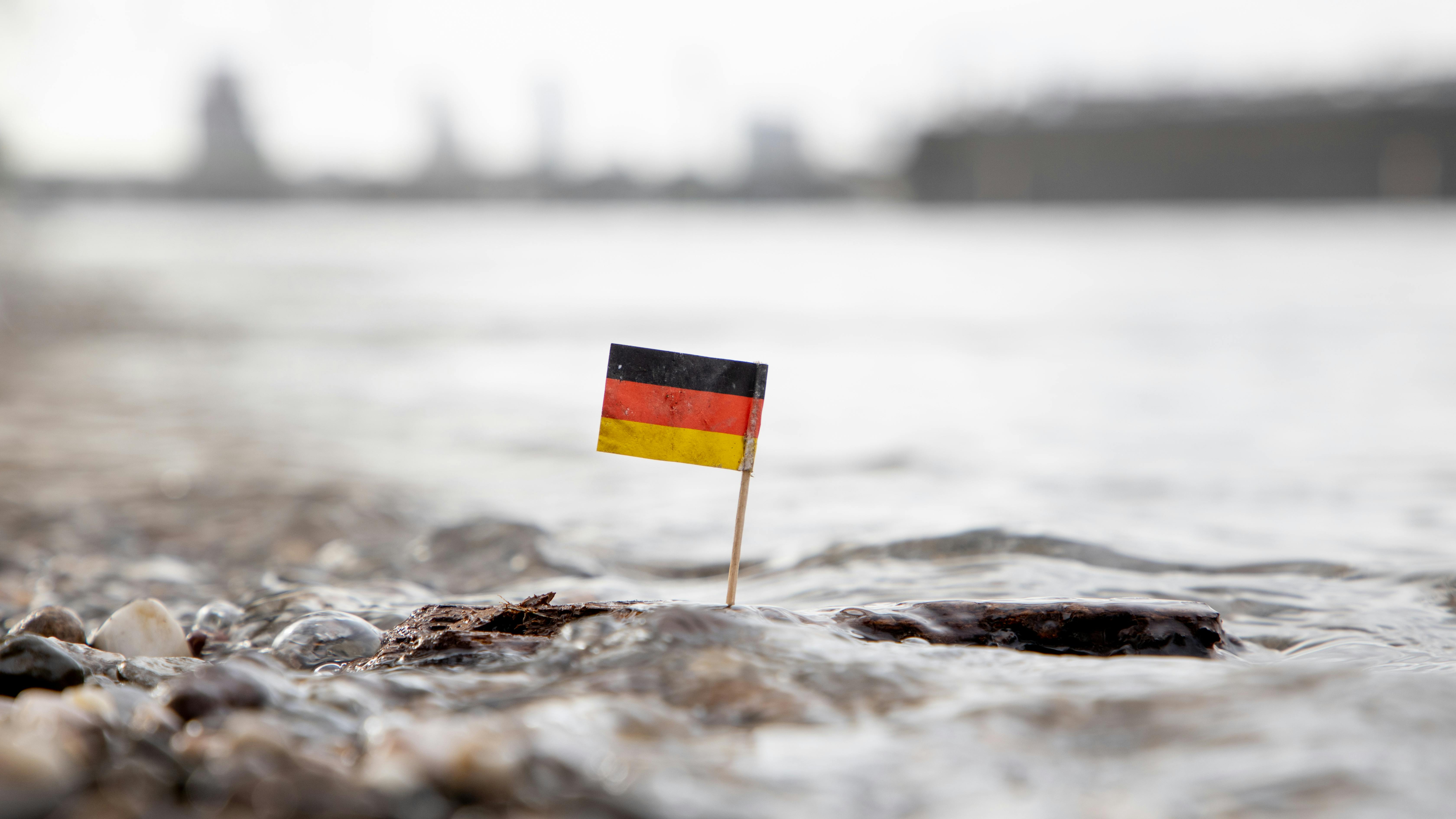 Eine einsame Deutschlandfahne treibt auf einem Stück Holz im Wasser, im Hintergrund die Silhouette einer Stadt.