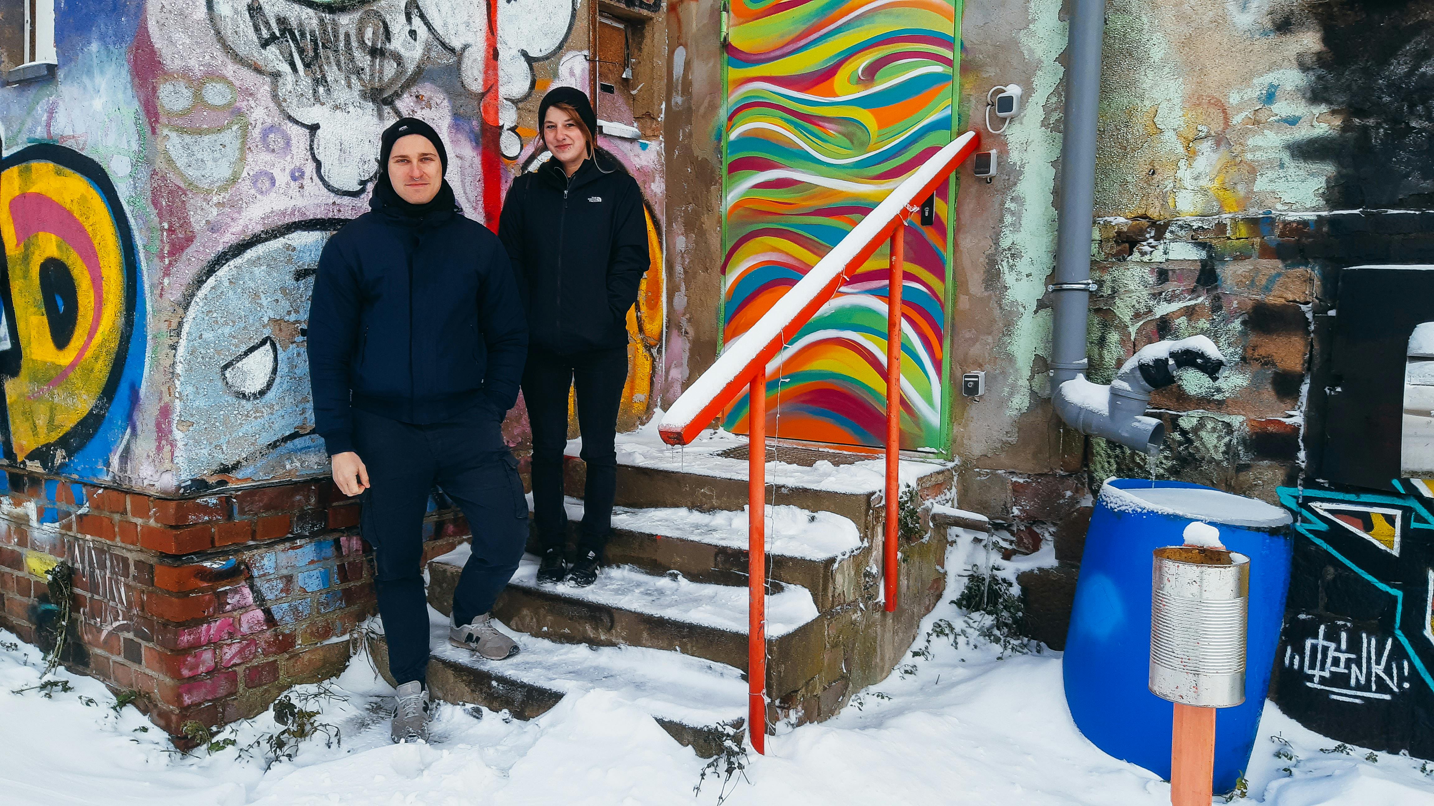 Elisa Grobe und Robert Weis stehen auf einer Treppe vor einen mit Graffiti bemalten Haus. Es ist Winter, sie tragen dunkle Winterklamotten. Beide haben Piercings im Gesicht.