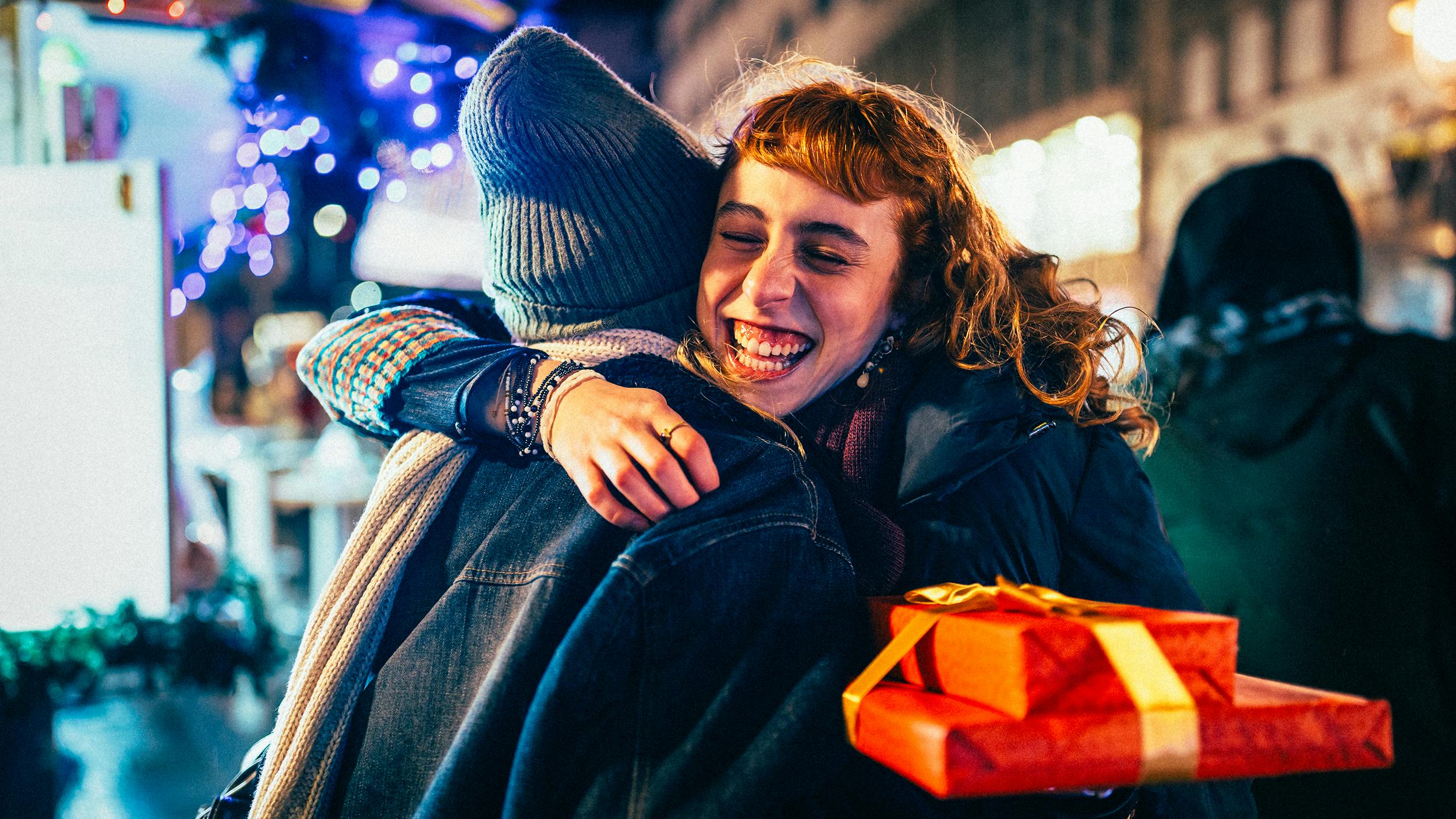 Eine Frau lacht herzlich, während sie eine andere Person umarmt. Die Frau hält ein Geschenk in der Hand.