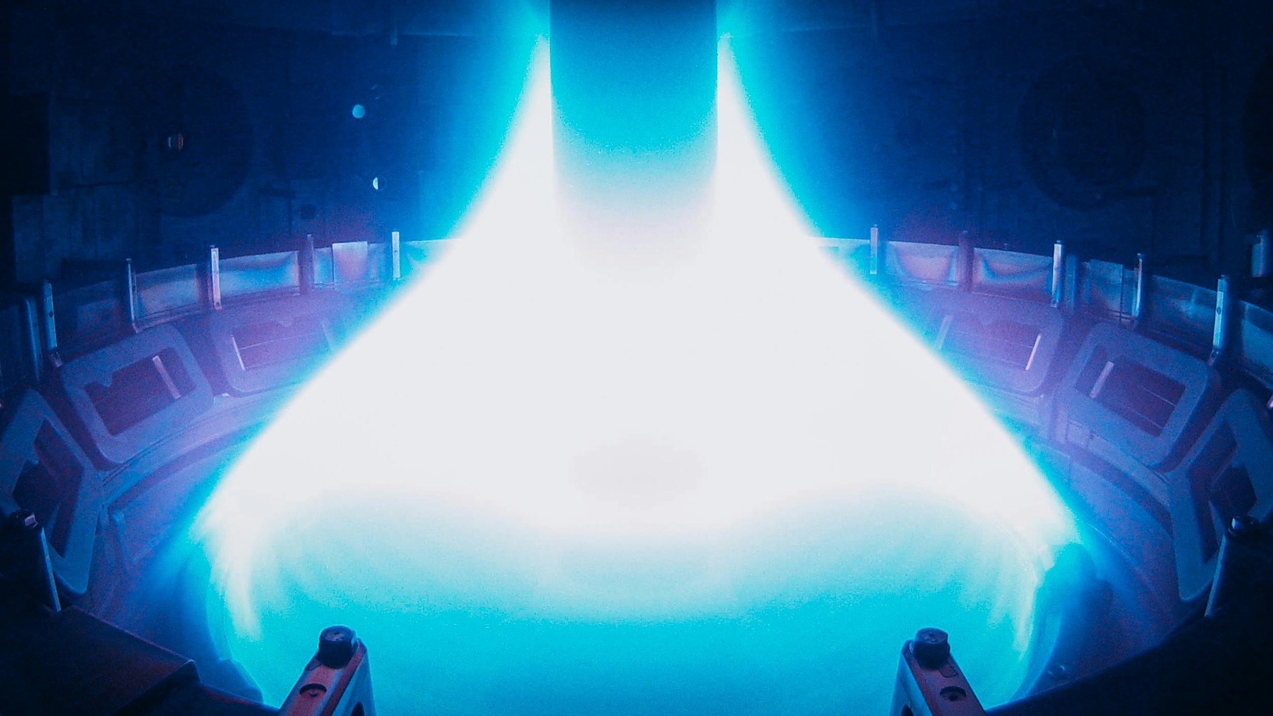 Foto vom Inneren eines Kernfusionsreaktors. Man sieht blau leuchtendes Plasma, das sich kegelförmig um eine senkrechte Metallstange bildet.