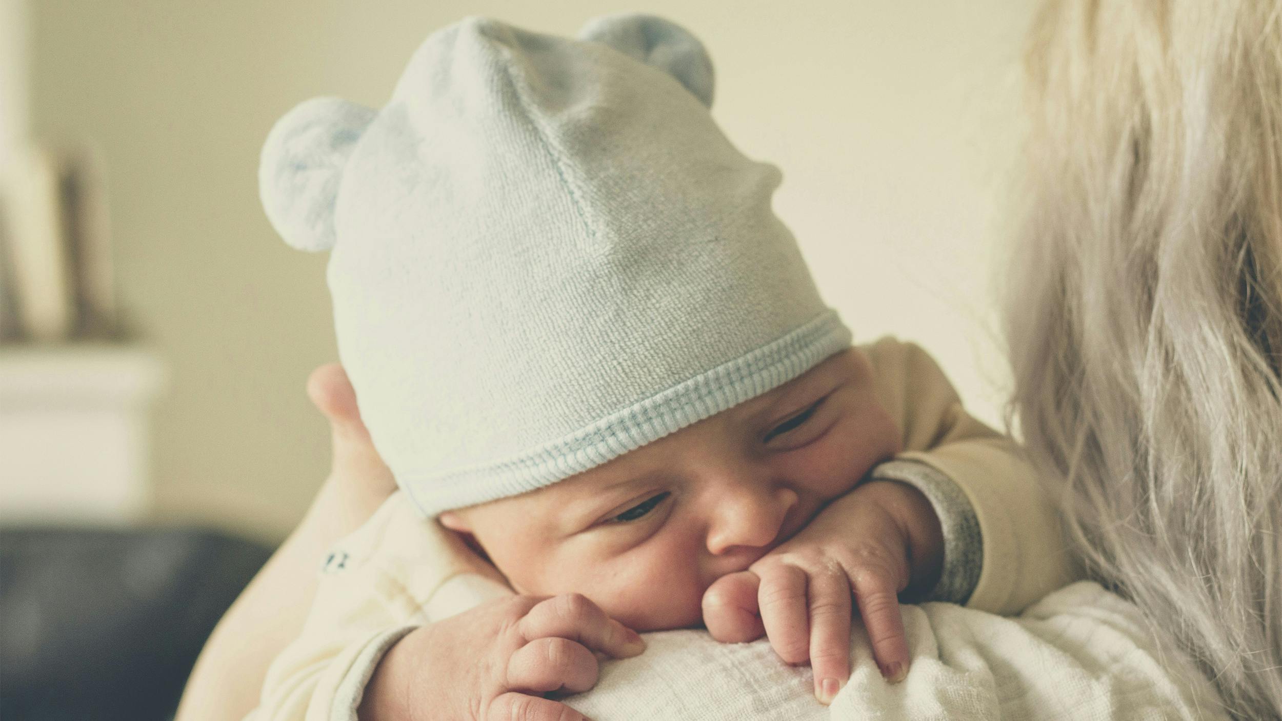 Ein Baby wird von einer älteren Frau auf dem Arm getragen. Es trägt eine hellblaue Mütze mit Bärenohren.