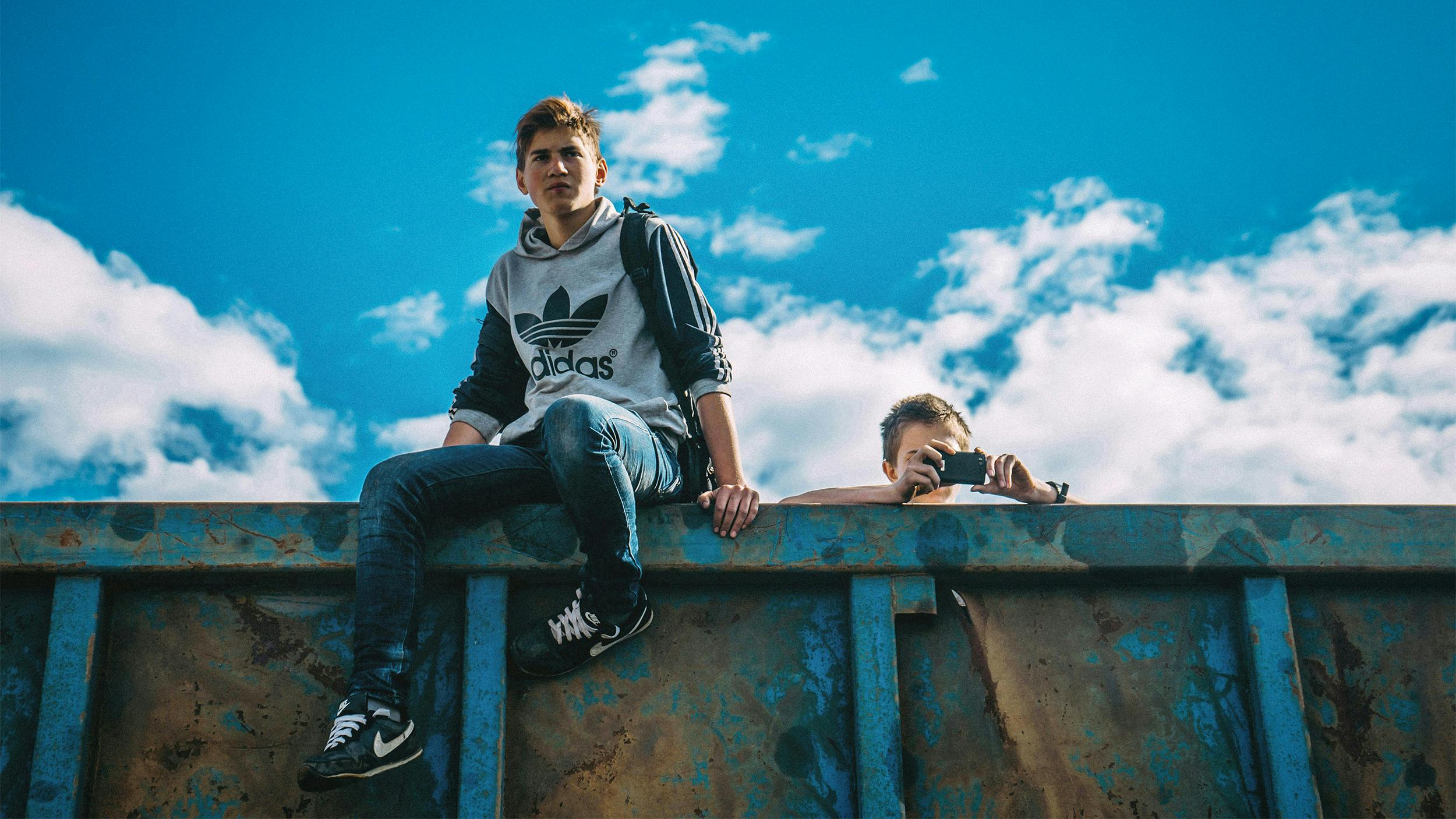 Ein Teenager-Junge sitzt auf einer Mauer. Hinter der Mauer steht ein zweiter Junge, man sieht nur seinen Kopf. Er macht gerade ein Foto mit seinem Handy.