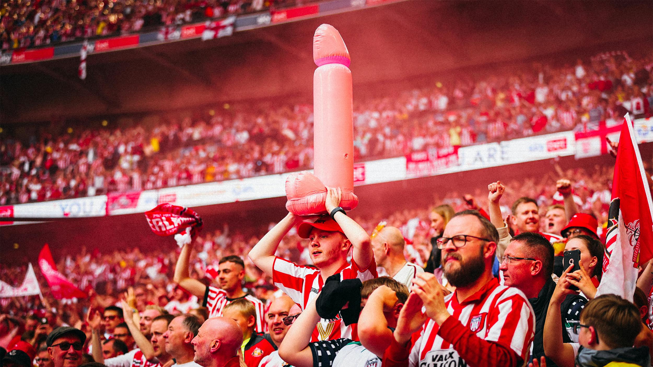 Fußballfans bei einem Spiel. Einer von ihnen hält einen aufblasbaren Penis über seinen Kopf.