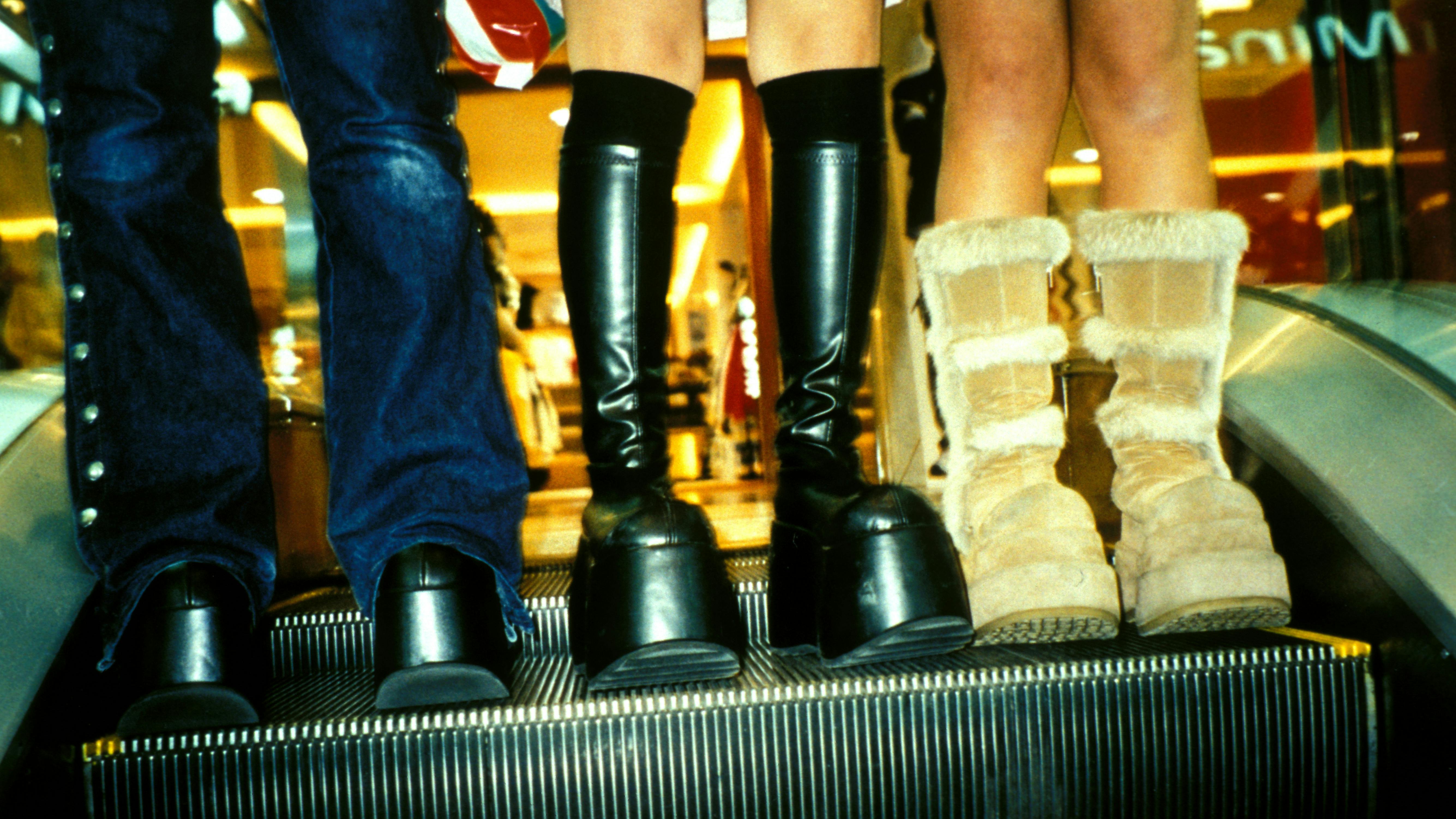 Drei junge Frauen stehen auf einer Rolltreppe. Man sieht nur ihre Stiefel.