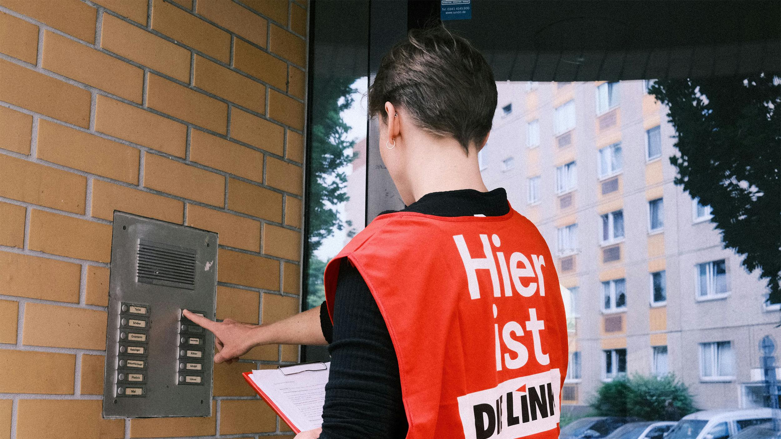 Eine Person mit einer roten Warnweste klingelt an einer Haustür.