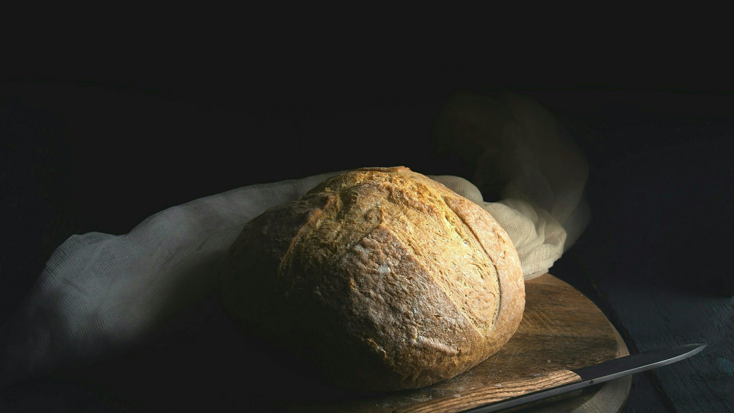 Ein Brot auf einem Brett, dahinter liegt weißer Stoff. Ein Messer liegt davor.