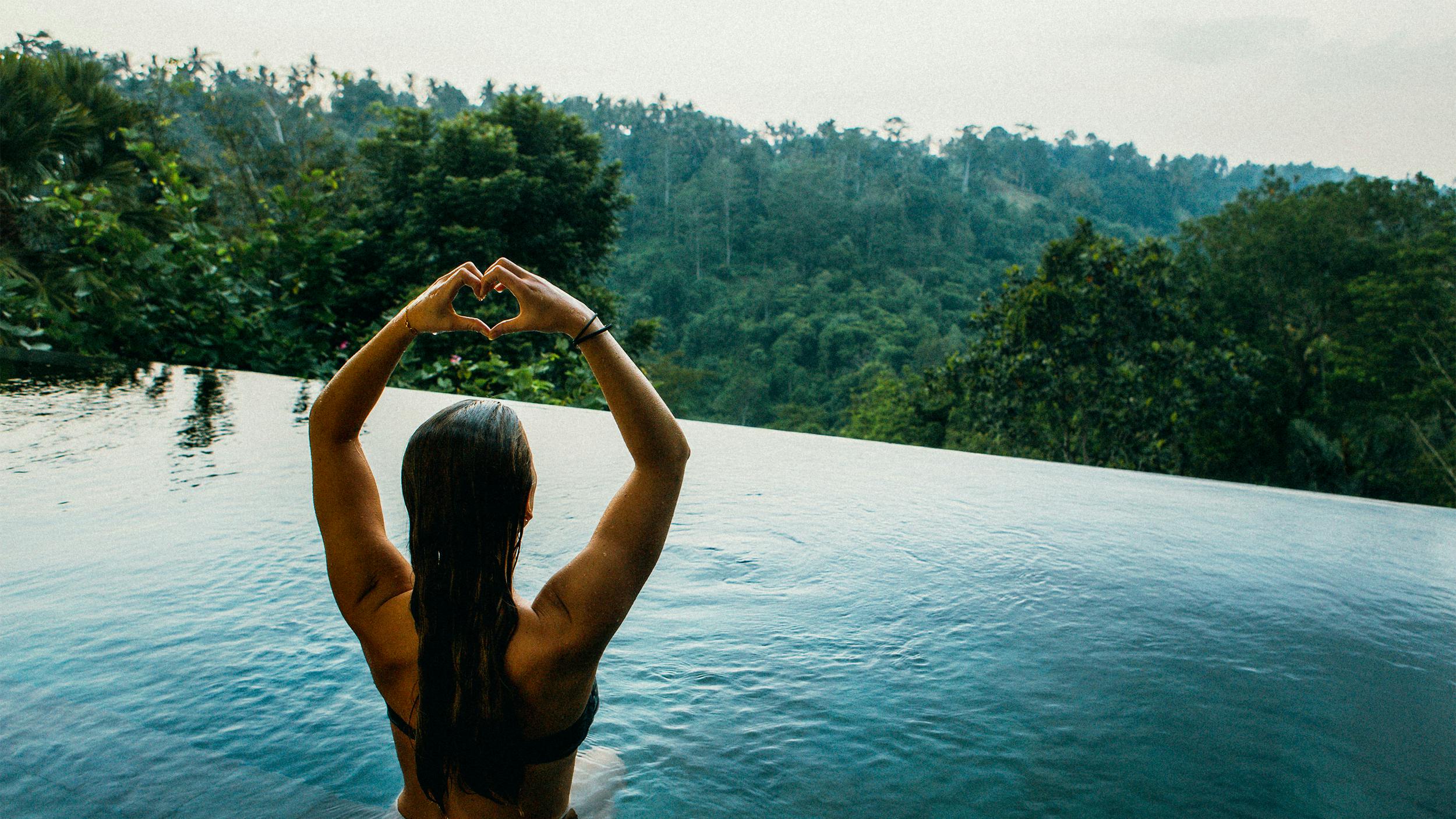 Eine Frau sitzt in einem Infinity Pool, von dem aus man auf einen Wald blicken kann. Sie hält die Arme über den Kopf und formt ihre Hände zu einem Herzen.