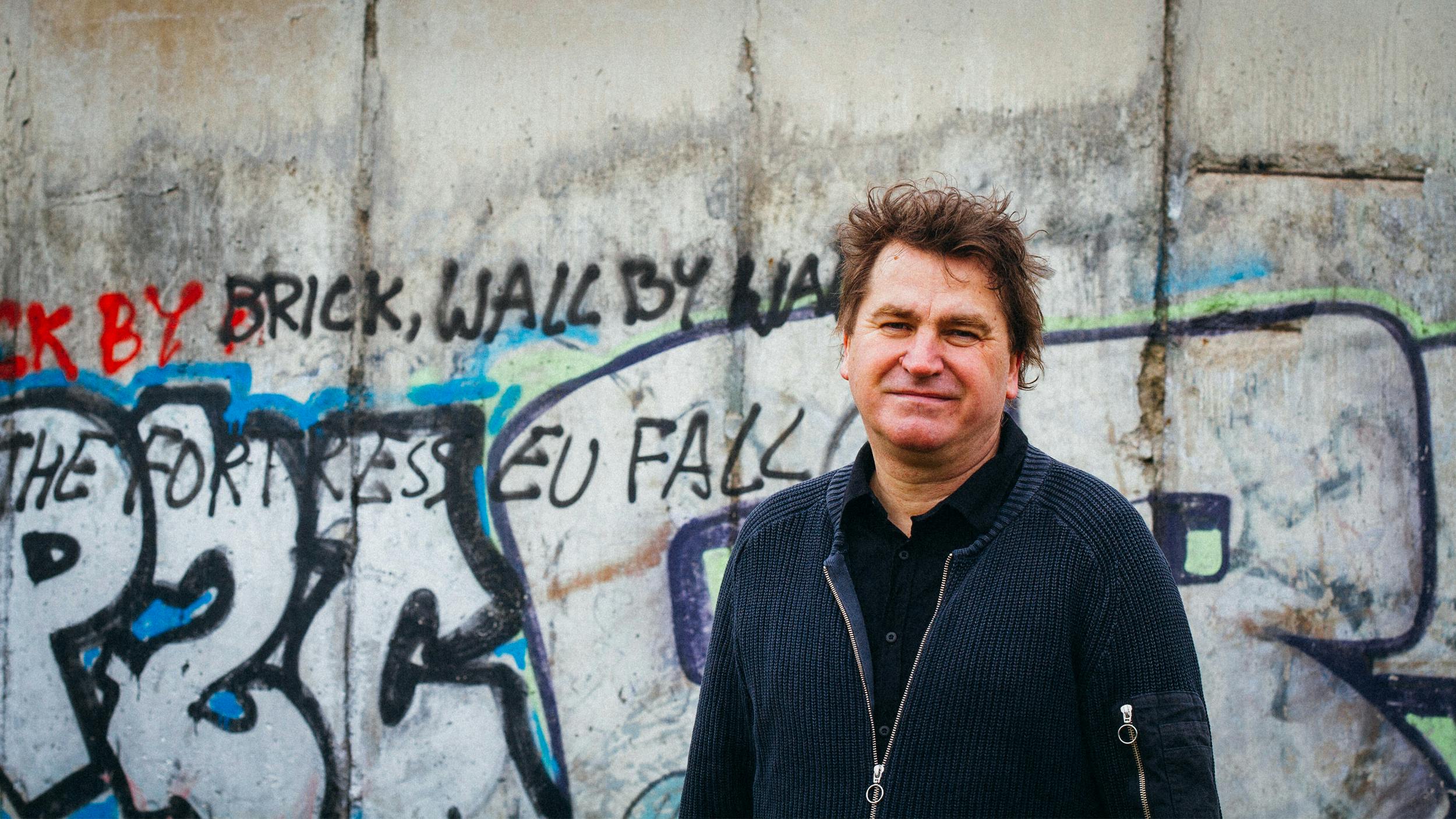 Ilko-Sascha Kowalczuk steht vor der ehemaligen Grenzmauer zwischen DDR und BRD. Sie ist mit Graffiti überzogen .Er blickt in die Kamera. 