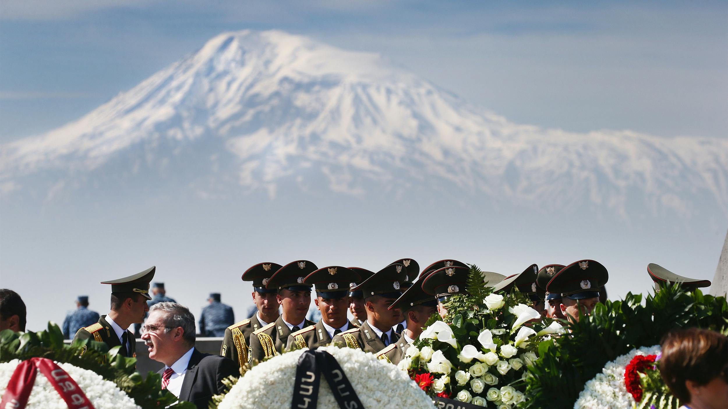 Soldaten in Uniform stehen vor Blumenkränzen die zum Gedenken aufgestellt wurden. Im Hintergrund sieht man einen Berg.