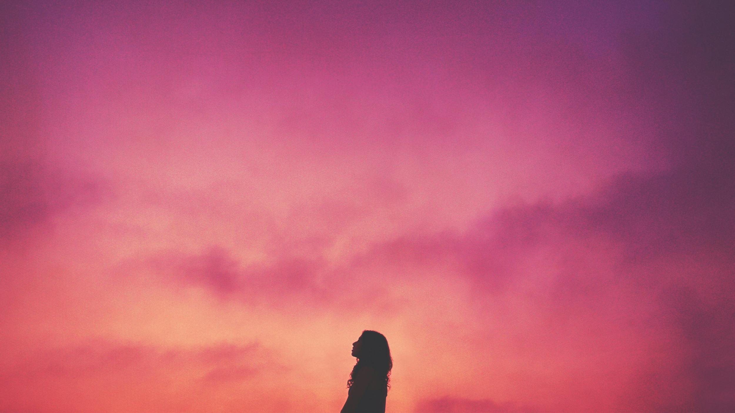 Eine Frau blickt in den Abendhimmel. Er ist in Rosa- und Orangetönen gefärbt.