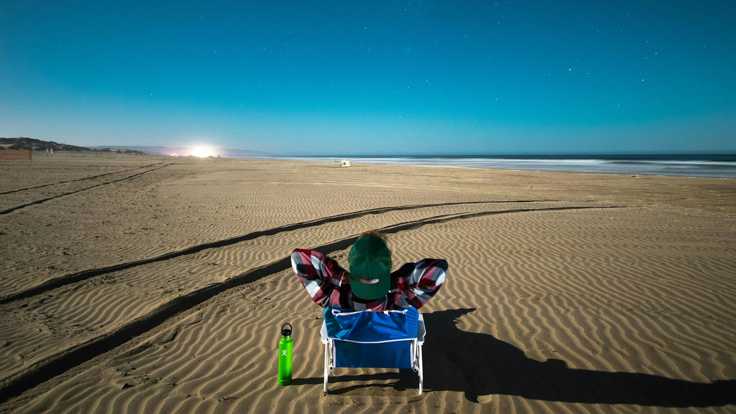 Ein Mann liegt in einer Liege am Strand und schaut auf das Meer. Am Himmel sind Sterne zu erkennen.