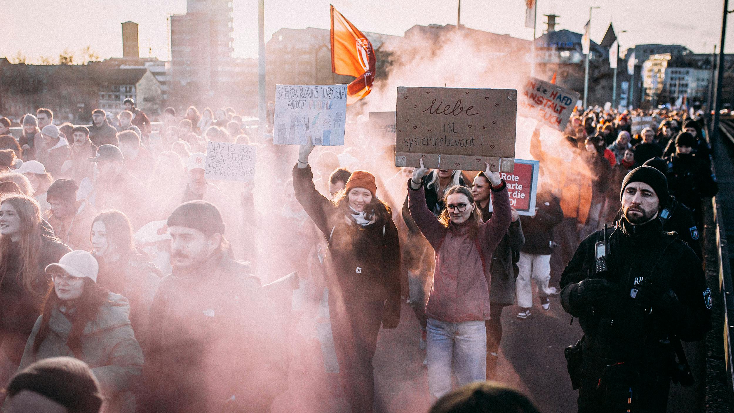 Junge Menschen laufen eine Straße entlang, sie halten Schilder in die Höhe. Im Hintergrund ziehen rosa Rauchschwaden auf.