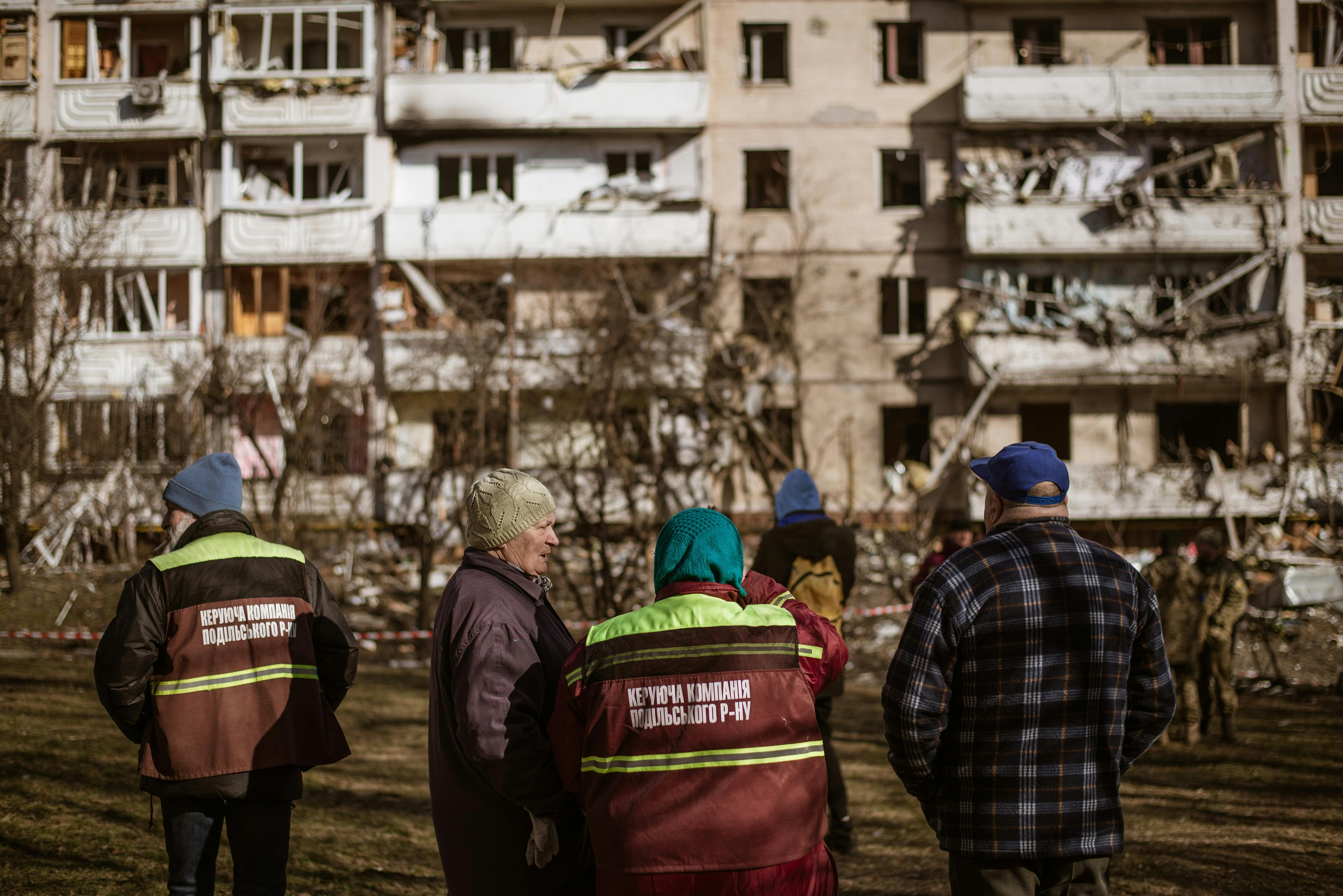 Menschen stehen vor einem zerbombten Wohnhaus in Kiev. Manche tragen Warnwesten.